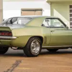 Seiten- und Heckansicht eines klassischen Chevrolet Camaro 350 V8 Coupé von 1969 mit grüner Metallic-Lackierung, geparktem Fahrzeug vor moderner Halle