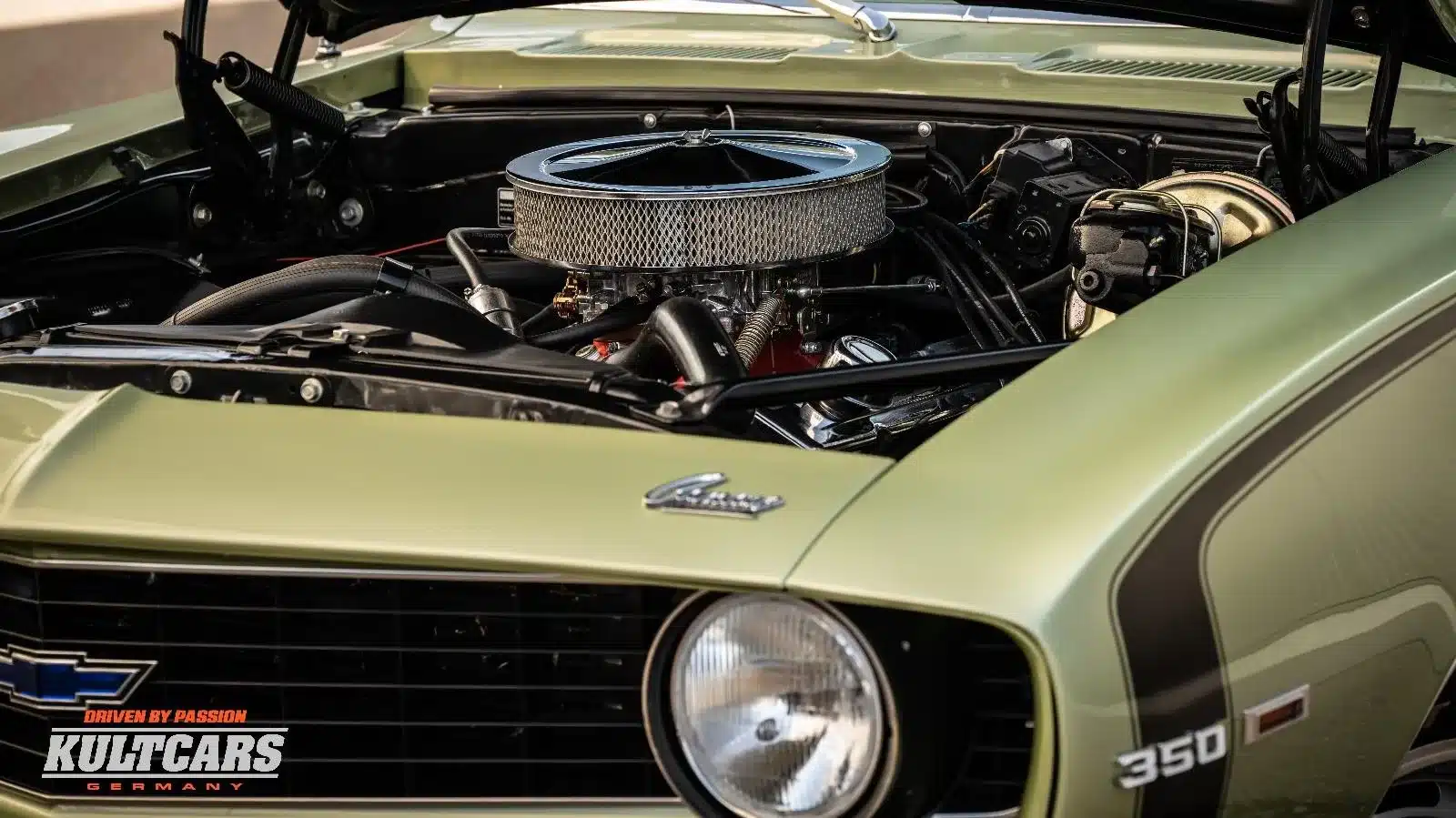 Nahaufnahme des Motorraums eines Chevrolet Camaro 350 V8 von 1969, Frost Green Lackierung, mit sichtbarem V8-Motor und Camaro-Emblem.