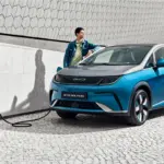 BYD Dolphin wird an einer Ladestation geladen, Mann lehnt sich an das Fahrzeug, modernes urbanes Umfeld.