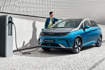 BYD Dolphin wird an einer Ladestation geladen, Mann lehnt sich an das Fahrzeug, modernes urbanes Umfeld.