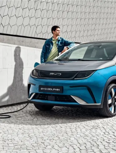 BYD Dolphin wird an einer Ladestation geladen, Mann lehnt sich an das Fahrzeug, modernes urbanes Umfeld.