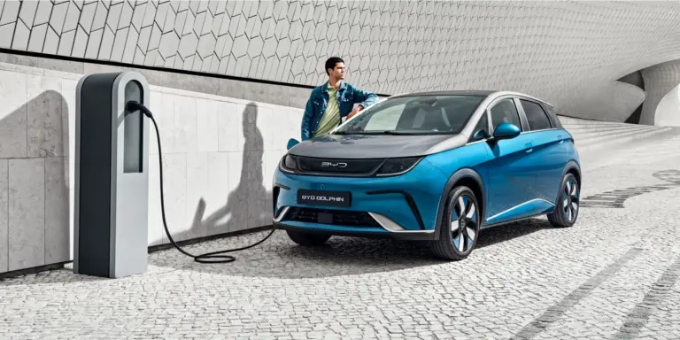 BYD Dolphin wird an einer Ladestation geladen, Mann lehnt sich an das Fahrzeug, modernes urbanes Umfeld.