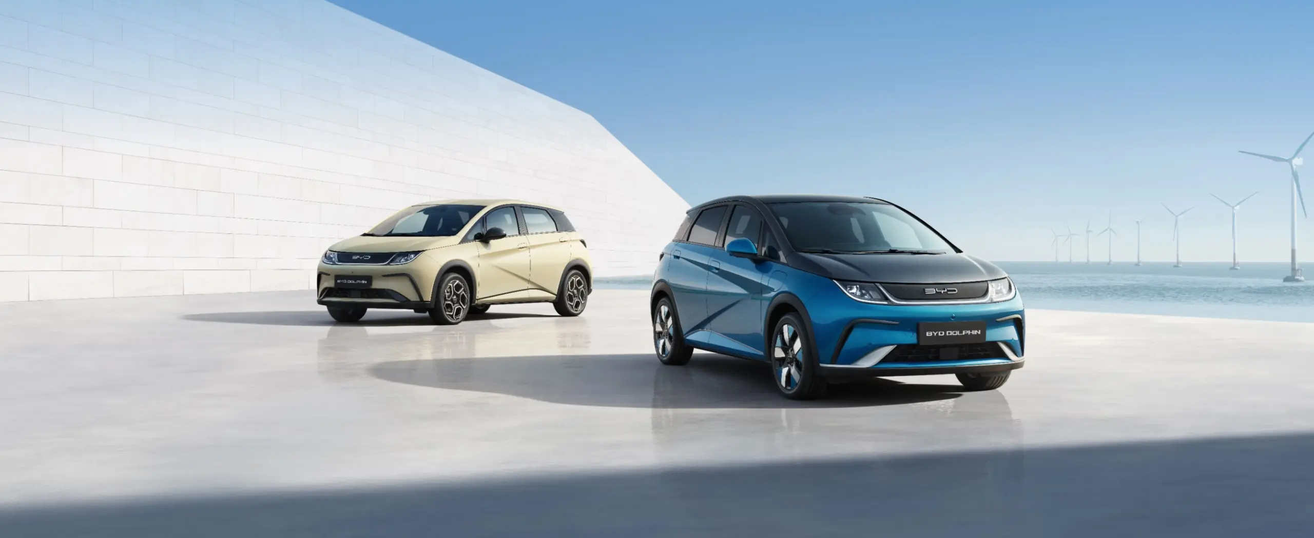 Zwei BYD Dolphin Elektroautos stehen auf einer hellen Fläche vor einer weißen Wand und dem Meer mit Windkraftanlagen im Hintergrund