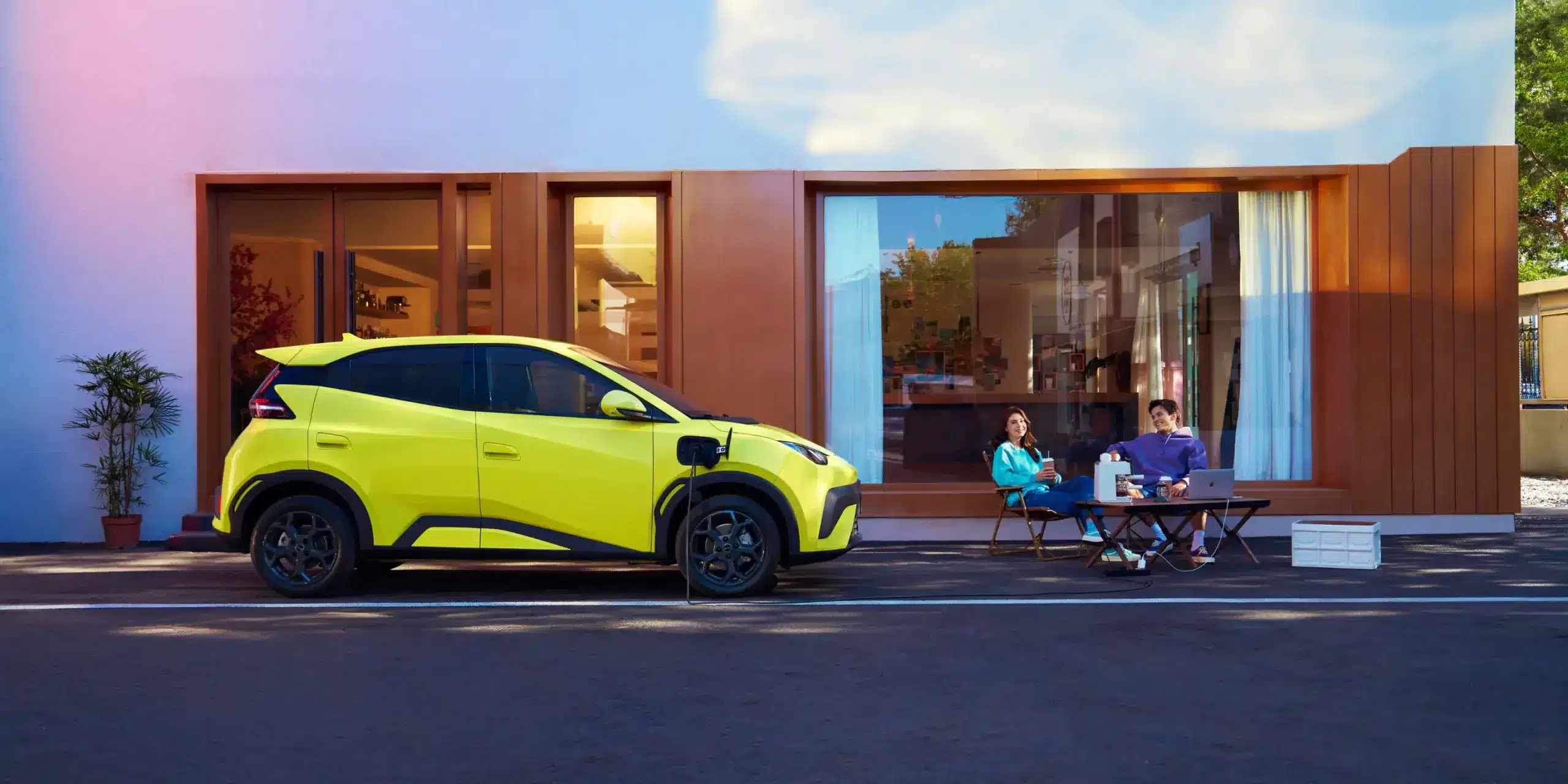 BYD Dolphin Surf in gelber Lackierung beim Laden vor einem Wohngebäude, daneben zwei Personen auf Klappstühlen.