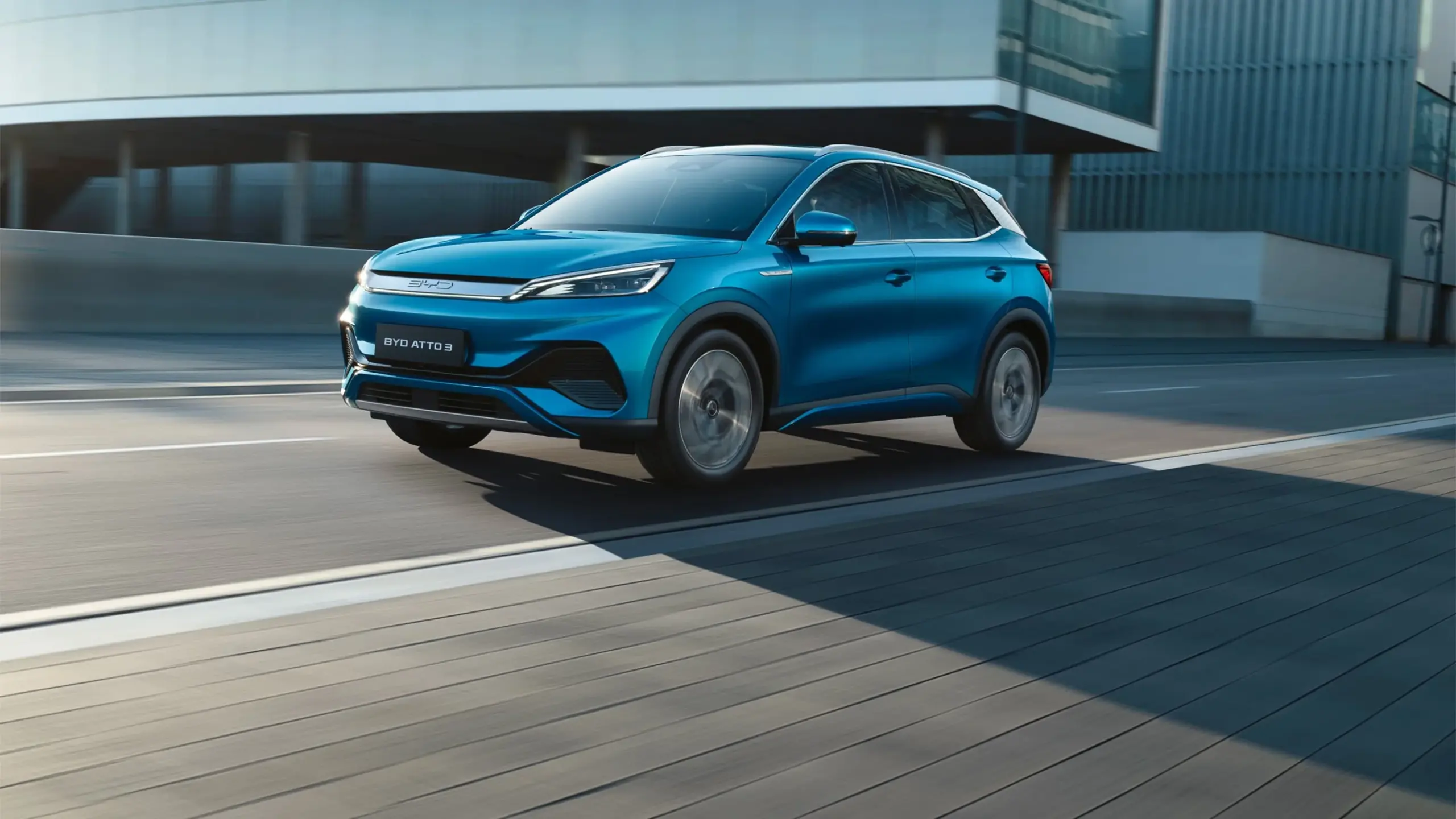Blauer BYD Atto 3 Elektro-SUV fährt auf einer Straße vor moderner Architektur.