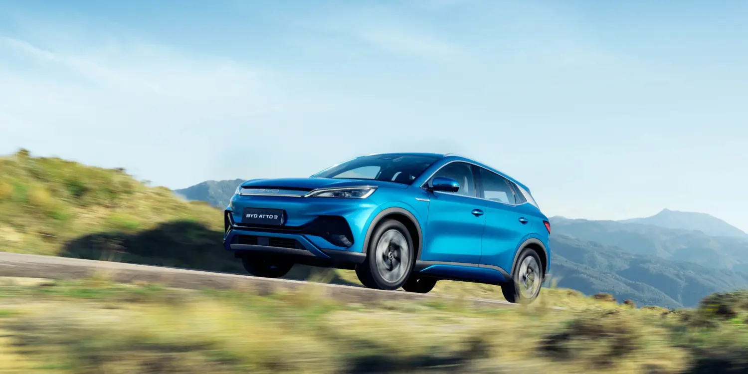 Blauer BYD Atto 3 Elektro-SUV fährt dynamisch auf einer Straße in landschaftlicher Umgebung mit Bergen im Hintergrund.
