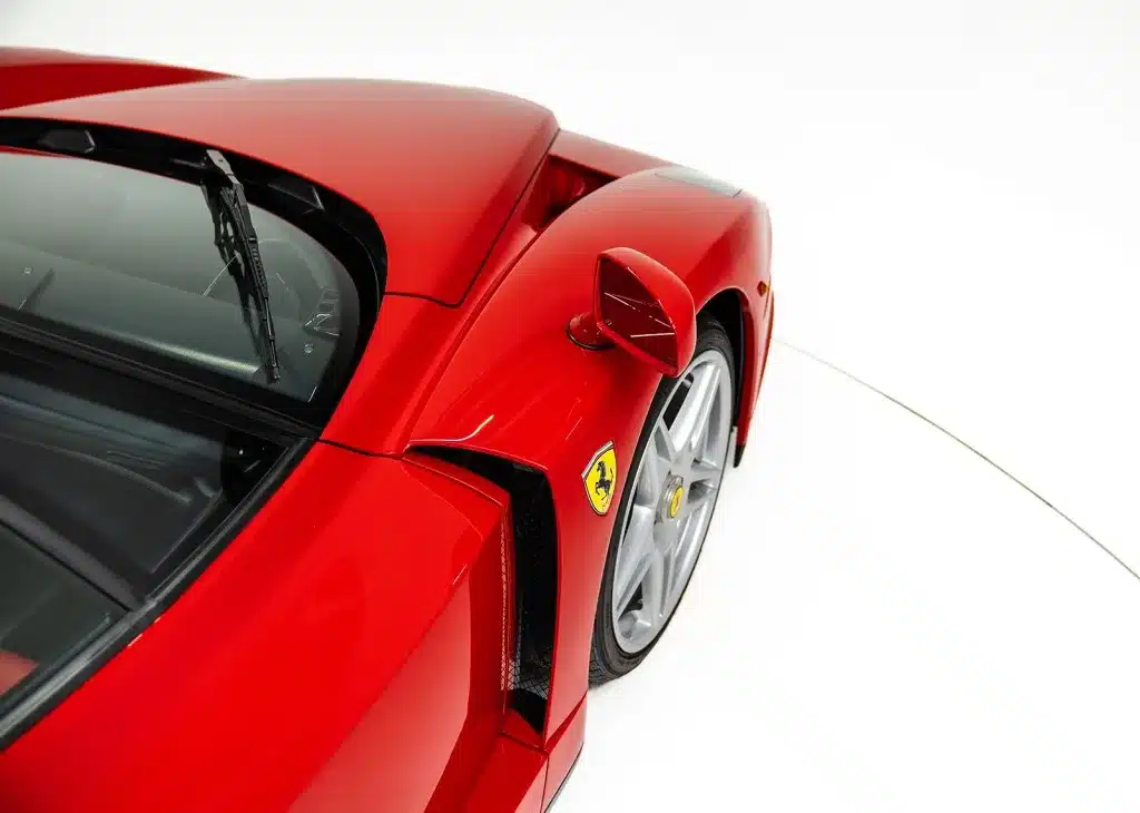 Detailaufnahme eines Ferrari Enzo in Rosso Corsa, mit Fokus auf Seitenspiegel, Hinterrad und das Ferrari-Logo.