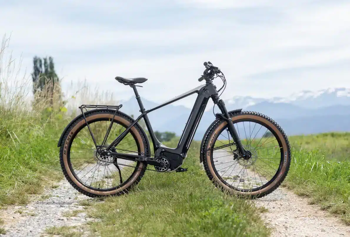 Schwarzes Heritage Style Escapade E-Tourenrad mit breiten Reifen und Gepäckträger, geparkt auf einem Feldweg mit bergiger Landschaft im Hintergrund.