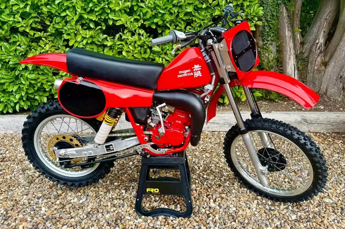 Honda CR 125 Mugen-Banzai-Umbau von 1980, seitliche Ansicht mit rotem Tank, Aluminium-Schwinge und Öhlins-Federbeinen, auf einem Ständer stehend.