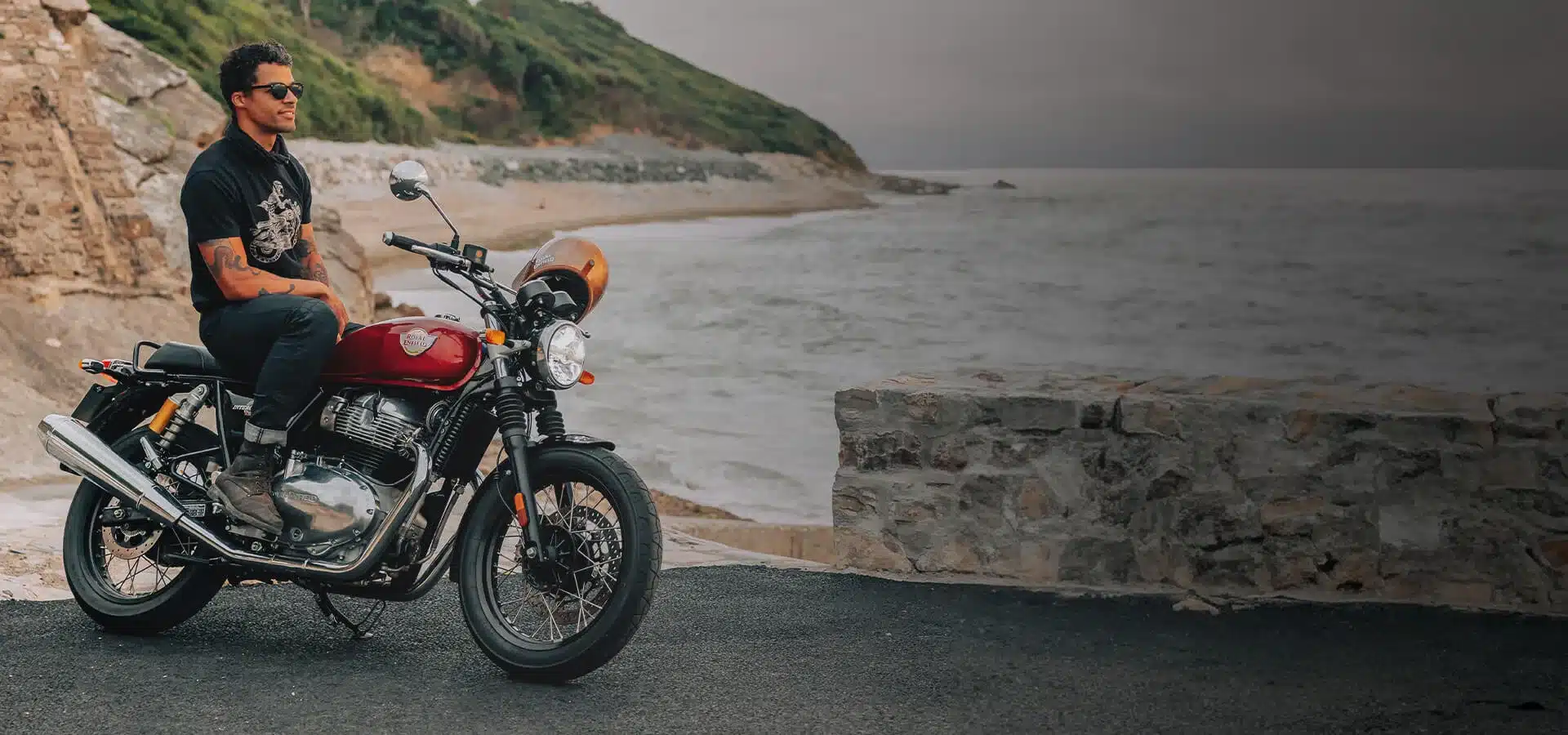 Mann sitzt auf einer Royal Enfield Interceptor 650 vor dem Meer und einer Felsenküste.