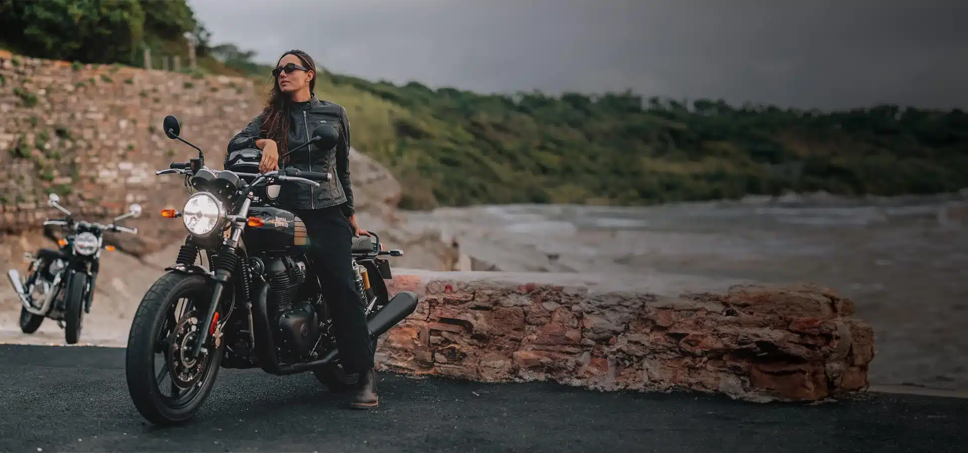 Royal Enfield Interceptor 650 als schwarzes Roadster-Motorrad mit Fahrerin an einer Steinmauer mit Meeresblick.