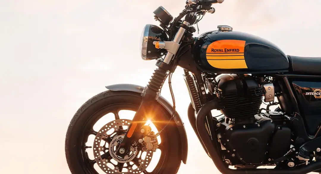 Royal Enfield Interceptor 650 Motorrad mit gelb-schwarzem Tank, seitlich fotografiert, Sonnenstrahlen im Vorderrad, Meer im Hintergrund.