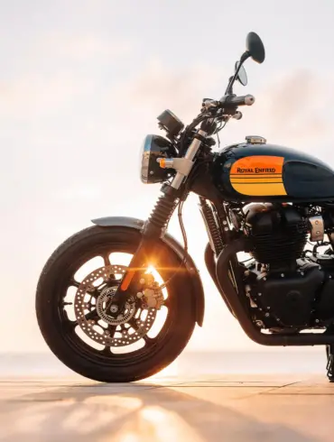 Royal Enfield Interceptor 650 Motorrad mit gelb-schwarzem Tank, seitlich fotografiert, Sonnenstrahlen im Vorderrad, Meer im Hintergrund.