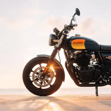 Royal Enfield Interceptor 650 Motorrad mit gelb-schwarzem Tank, seitlich fotografiert, Sonnenstrahlen im Vorderrad, Meer im Hintergrund.