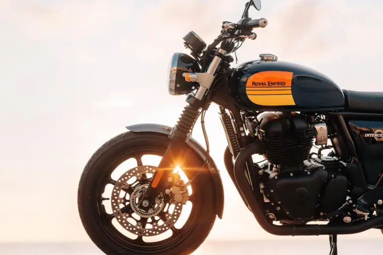 Royal Enfield Interceptor 650 Motorrad mit gelb-schwarzem Tank, seitlich fotografiert, Sonnenstrahlen im Vorderrad, Meer im Hintergrund.