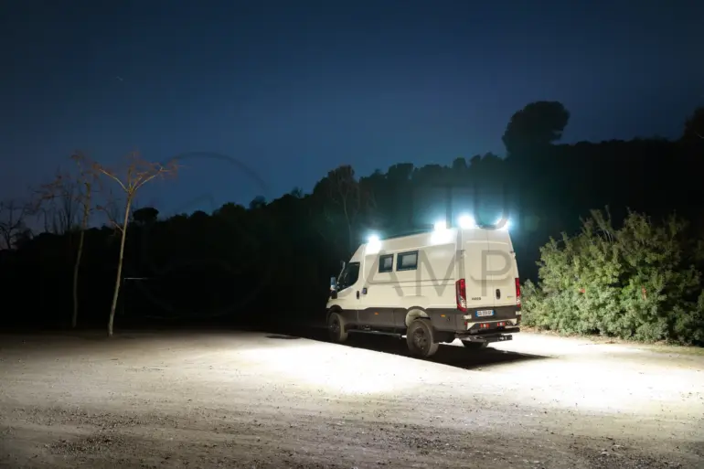 Iveco Daily 4x4 L3H2 Camper mit eingeschalteter Außenbeleuchtung bei Nacht auf offenem Gelände