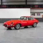 Ein rotes Jaguar E-Type Coupé Serie 1 steht in einer modernen Halle auf grauen Fliesen.