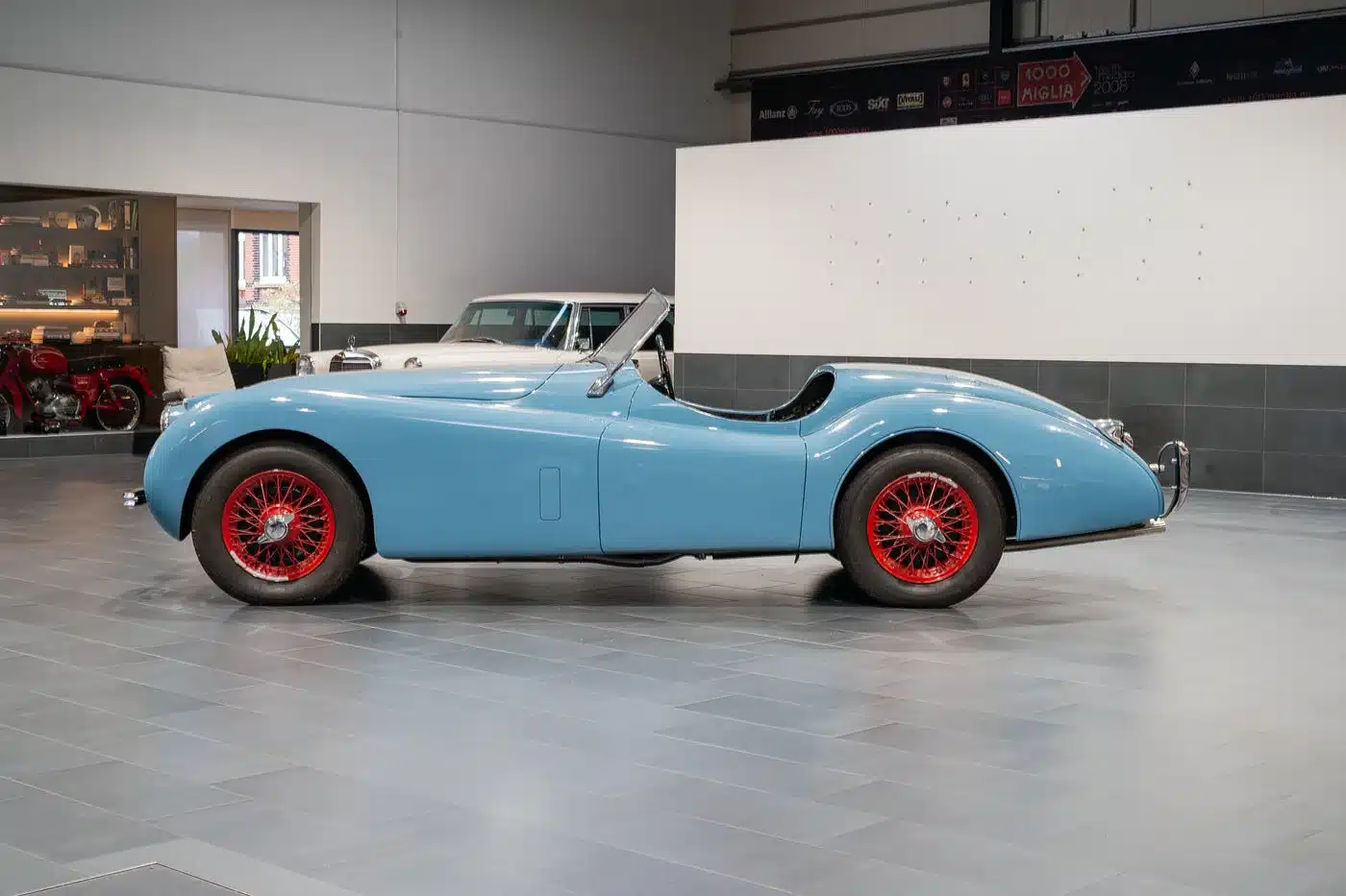 Seitenansicht einer klassischen Jaguar Karosserie in Blau mit roten Speichenrädern in einer Halle.