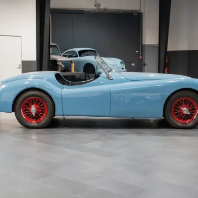 Seitenansicht einer blauen Jaguar Karosserie mit roten Speichenrädern in einer modernen Werkstatt