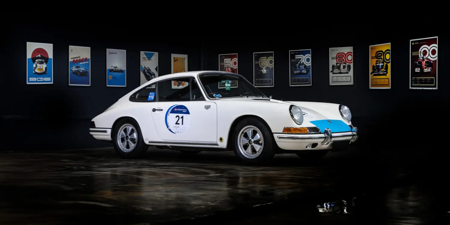 Seitenansicht eines weißen klassischen Sportwagens mit Startnummer, ausgestellt vor Motorsport-Postern.
