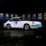 Seitenansicht eines weißen klassischen Sportwagens mit Startnummer, ausgestellt vor Motorsport-Postern.