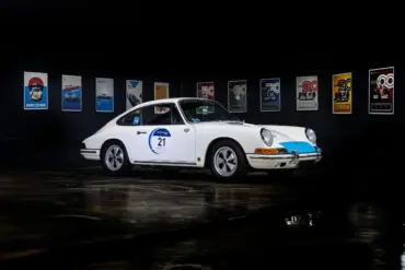 Seitenansicht eines weißen klassischen Sportwagens mit Startnummer, ausgestellt vor Motorsport-Postern.