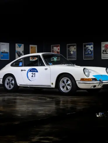 Seitenansicht eines weißen klassischen Sportwagens mit Startnummer, ausgestellt vor Motorsport-Postern.