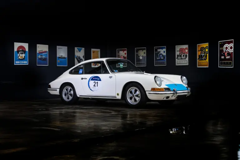 Seitenansicht eines weißen klassischen Sportwagens mit Startnummer, ausgestellt vor Motorsport-Postern.