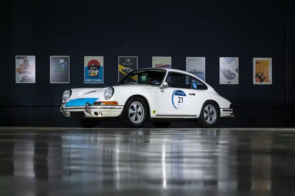 Weißer Oldtimer-Coupé, vermutlich Porsche 911, in einer modernen Halle mit Poster im Hintergrund.