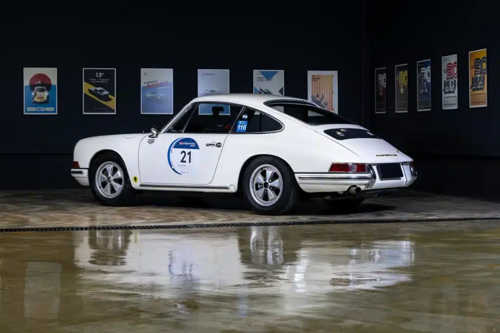 Seitenansicht eines weißen klassischen Porsche 911 Coupés mit Startnummer, ausgestellt in einer modernen Halle vor Automobilpostern.