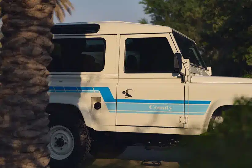 Land Rover 90 County von 1985 in Elfenbeinweiß mit blauem Streifendesign, fotografiert im Sonnenlicht von der Seite.