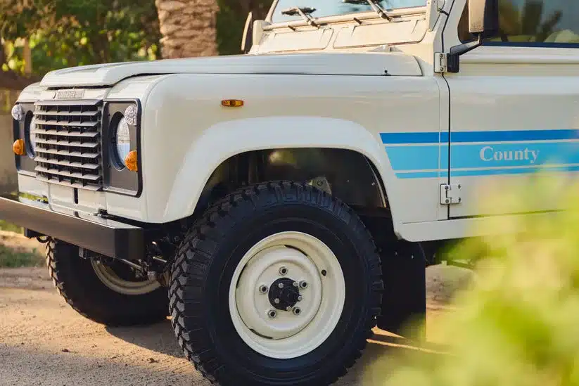 Front- und Seitenansicht eines restaurierten Land Rover 90 County, Baujahr 1985, in Ivory White mit blauen Streifen und robusten Geländereifen.