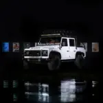 Weißer Land Rover Defender mit Offroad-Reifen im Studio, reflektiert auf glänzendem Boden.