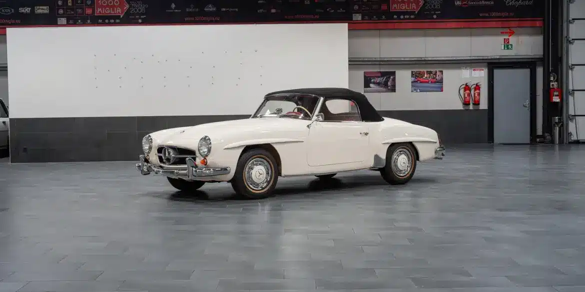 Seitenansicht eines weißen Mercedes-Benz 190 SL Roadster mit schwarzem Verdeck, Baujahr 1961, stehend in einer hellen Halle.
