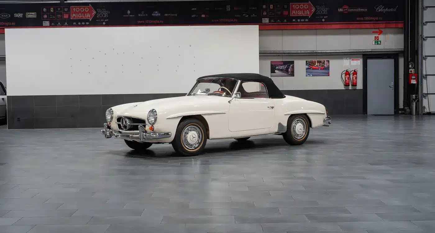 Seitenansicht eines weißen Mercedes-Benz 190 SL Roadster mit schwarzem Verdeck, Baujahr 1961, stehend in einer hellen Halle.