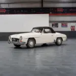 Seitenansicht eines weißen Mercedes-Benz 190 SL Roadster mit schwarzem Verdeck, Baujahr 1961, stehend in einer hellen Halle.