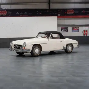 Seitenansicht eines weißen Mercedes-Benz 190 SL Roadster mit schwarzem Verdeck, Baujahr 1961, stehend in einer hellen Halle.