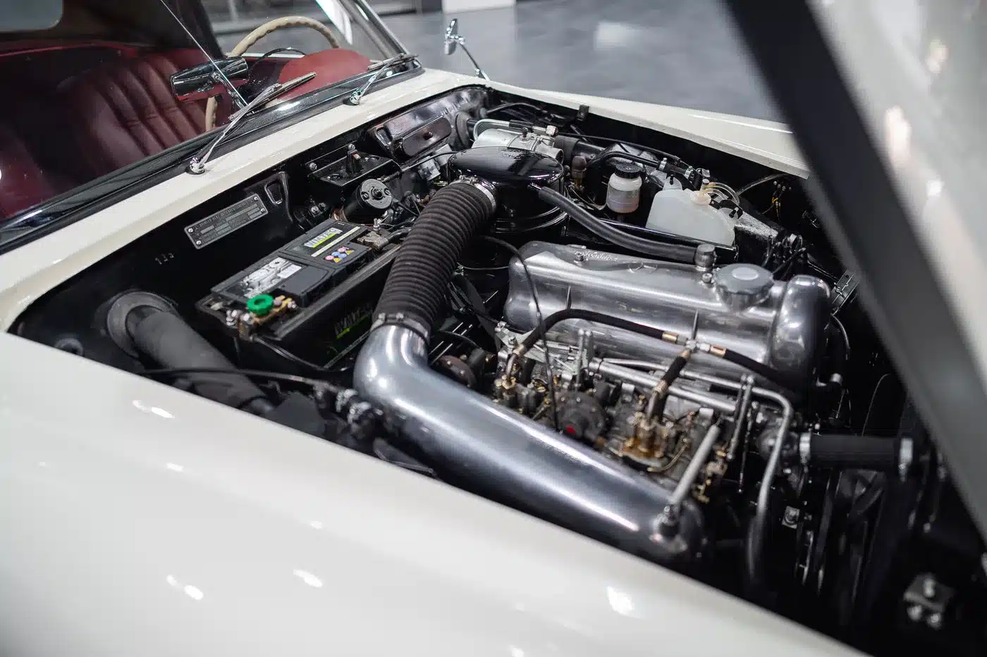 Geöffnete Motorhaube eines weißen Mercedes-Benz 190 SL, die den restaurierten Reihenvierzylinder-Motor und das rote Interieur zeigt.