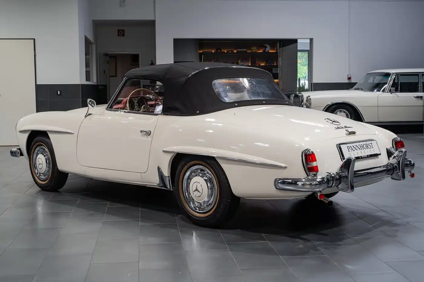 Seiten- und Heckansicht eines weißen Mercedes-Benz 190 SL (W121 B II) Baujahr 1961 mit schwarzem Verdeck in einer Ausstellungshalle.