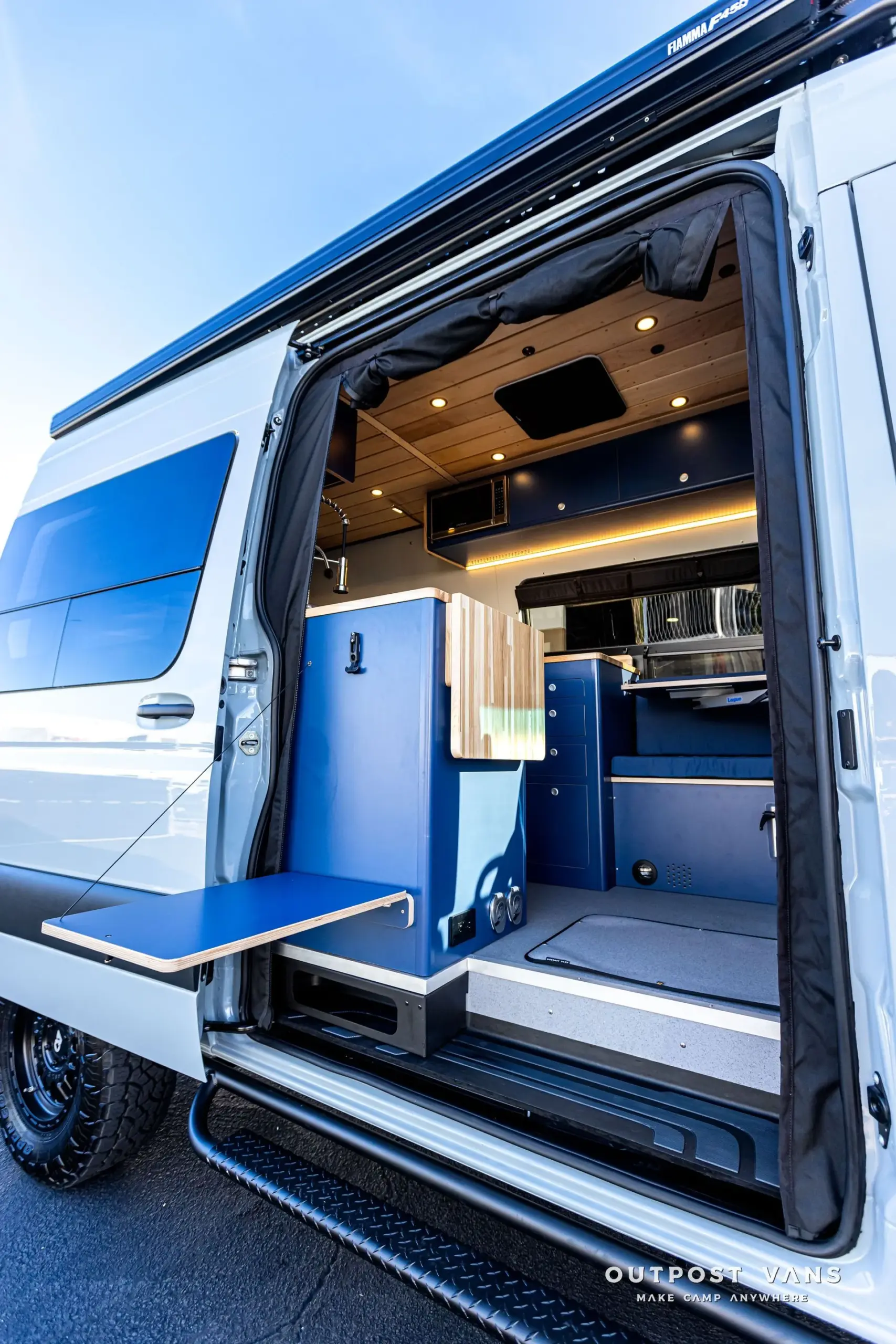 Innenraum eines Mercedes Sprinter Campers mit blauer Küchenzeile, Butcherblock-Arbeitsplatte und viel Stauraum, aufgenommen durch die seitliche Schiebetür.