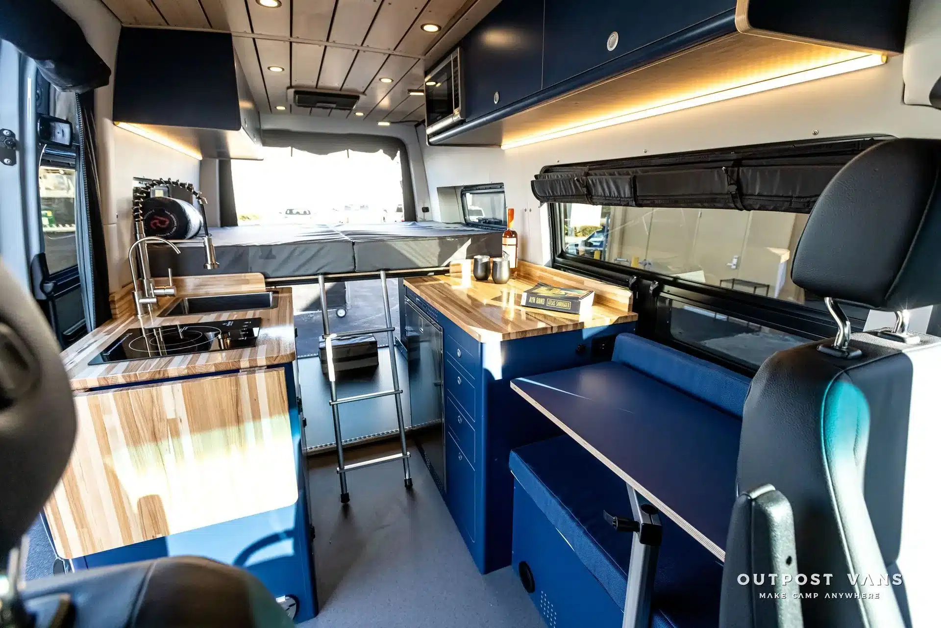 Innenansicht des Mercedes Sprinter El Duque mit Holzarbeitsflächen, Induktionskochfeld, Stauraum und Schlafbereich.