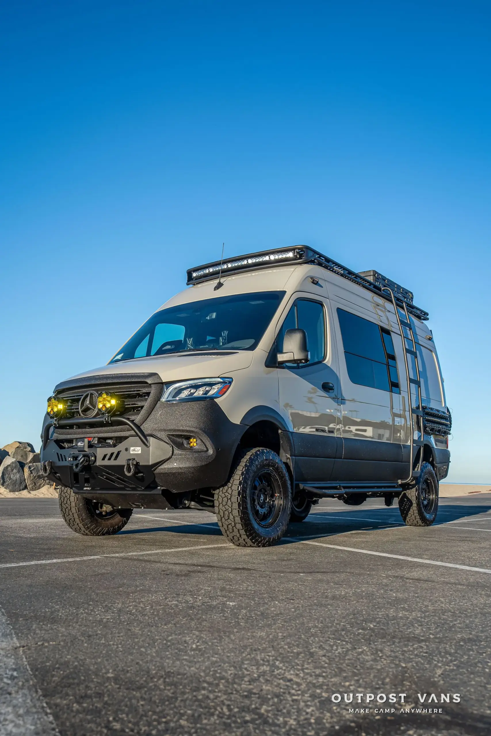 Mercedes-Benz Sprinter Lobo Allrad-Camper mit Dachträger, Leiter und Offroad-Ausstattung auf Asphaltfläche bei klarem Himmel.