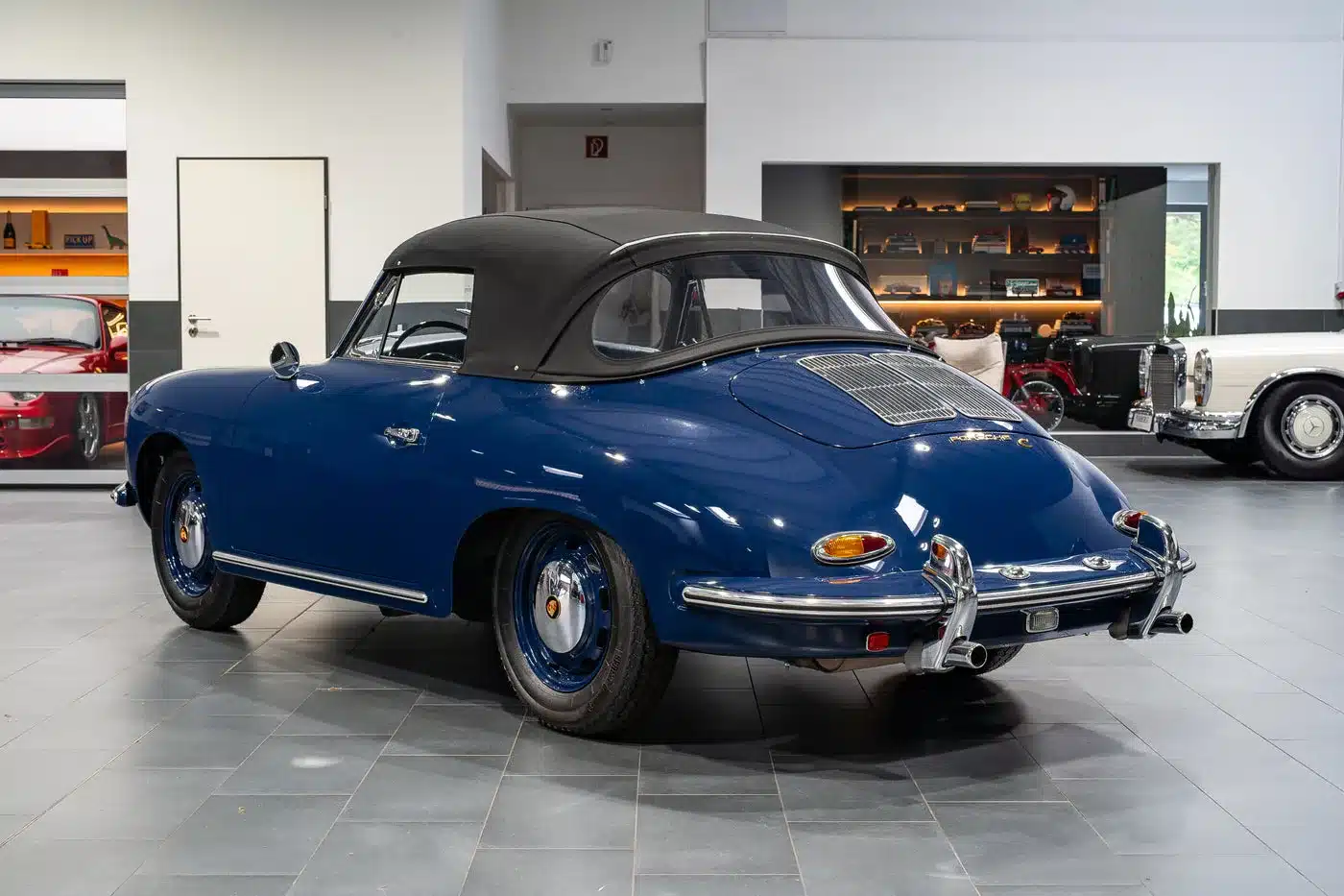 Blaues Porsche 356 C Cabriolet von 1964 mit geschlossenem Verdeck, fotografiert von schräg hinten in einer modernen Fahrzeughalle.
