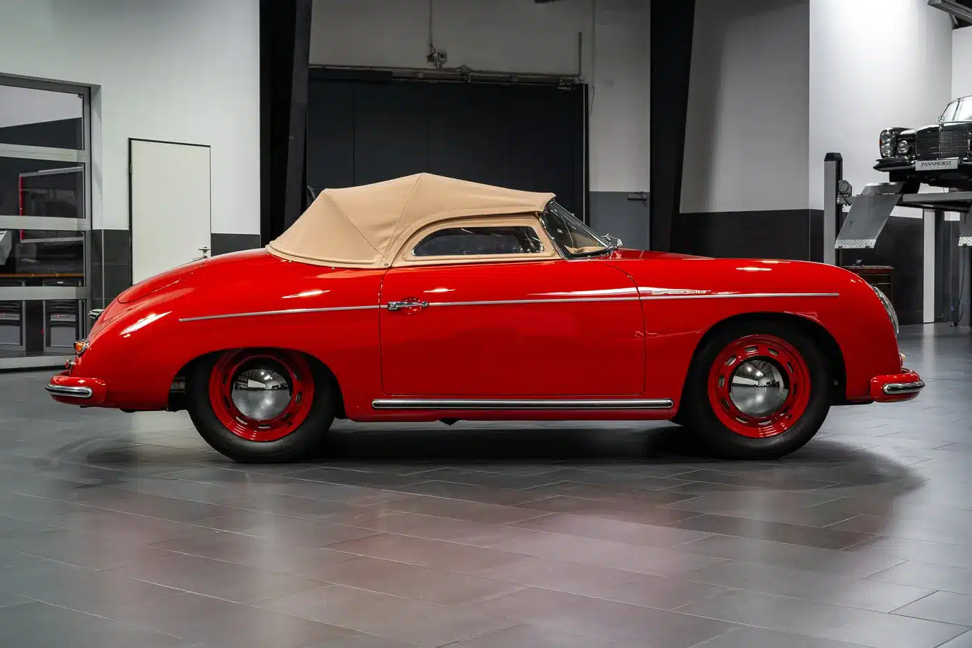 Seitenansicht eines roten Porsche 356 Pre-A Speedster von 1955 mit beigem Stoffverdeck, restauriert und in einer Werkstatt fotografiert.