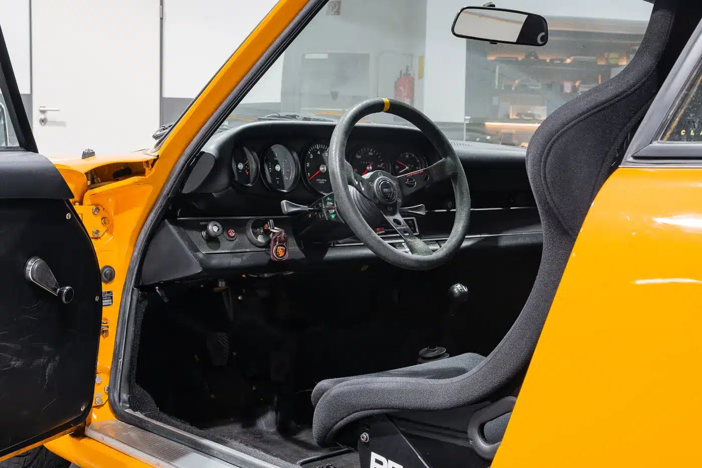 Innenraum eines klassischen Porsche 911 mit geöffnetem Fahrerfenster, schwarzem Rennsitz und sichtbaren Instrumenten.