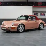 Porsche 964 Carrera 4 Cabriolet in roségold mit rotem Verdeck, seitlich fotografiert, in einer modernen Ausstellungshalle