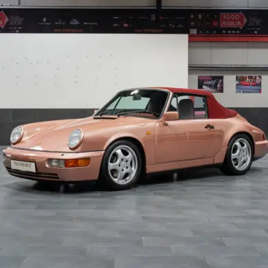 Porsche 964 Carrera 4 Cabriolet in roségold mit rotem Verdeck, seitlich fotografiert, in einer modernen Ausstellungshalle