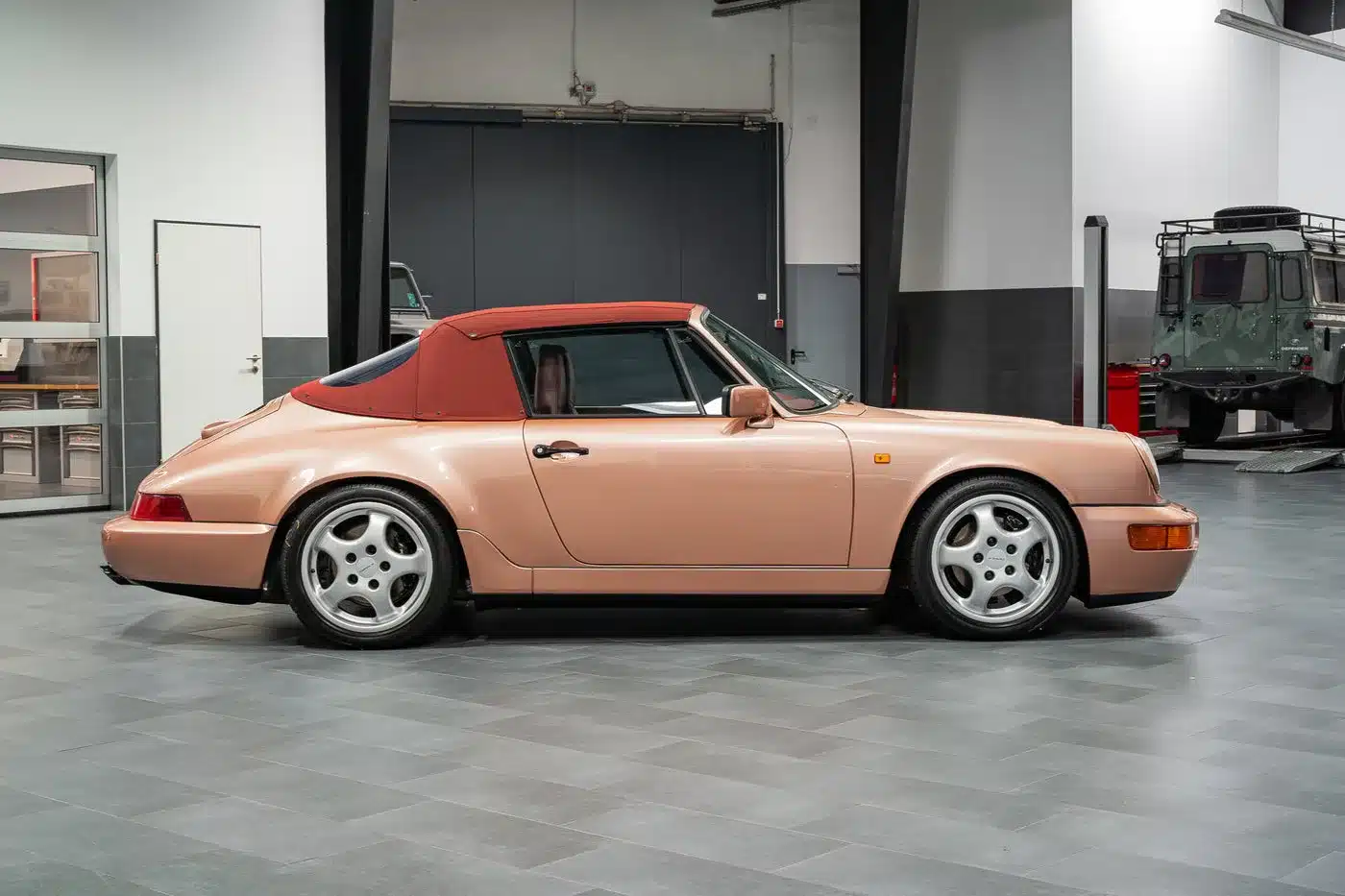 Porsche 964 Carrera 4 Cabriolet, Baujahr 1990–1993, in Roségold mit rotem Verdeck als Seitenansicht in einer Werkstatthalle