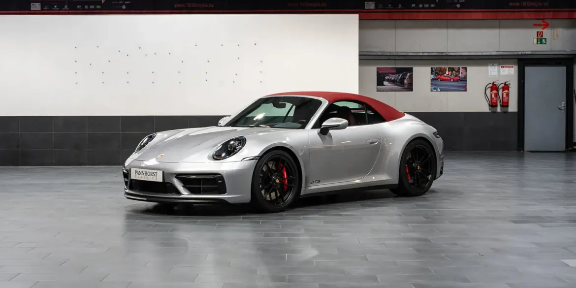 Porsche 992 911 Carrera GTS Cabriolet von 2023 in Silber mit geschlossenem rotem Verdeck, seitliche Frontansicht in einer Ausstellungshalle.