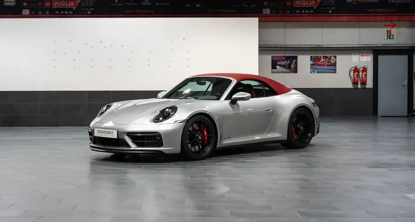 Porsche 992 911 Carrera GTS Cabriolet von 2023 in Silber mit geschlossenem rotem Verdeck, seitliche Frontansicht in einer Ausstellungshalle.